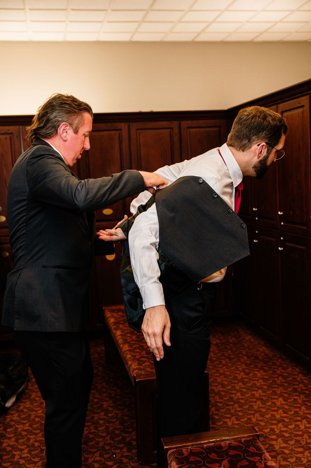 A man helps another put on a black vest in a wood-paneled dressing room, both wearing formal attire with red ties.