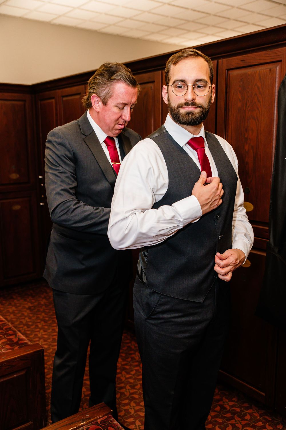 Two men in dark suits and red ties, one helping the other adjust his vest before a wedding ceremony.
