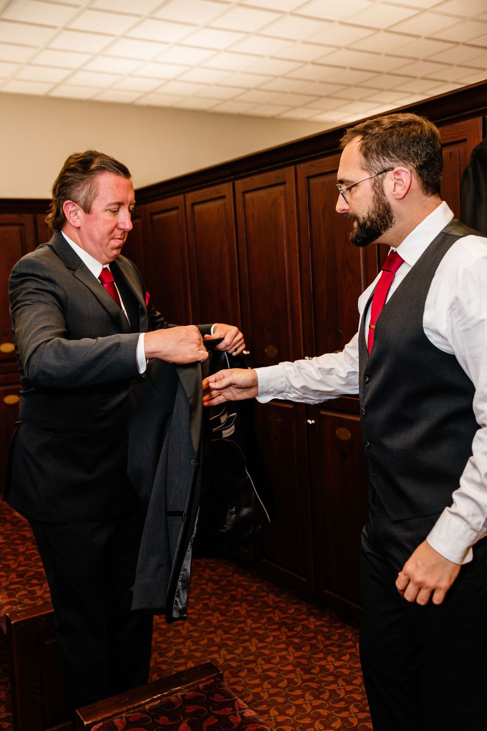 Two men in formal suits with red ties assist each other getting dressed in a wood-paneled room.