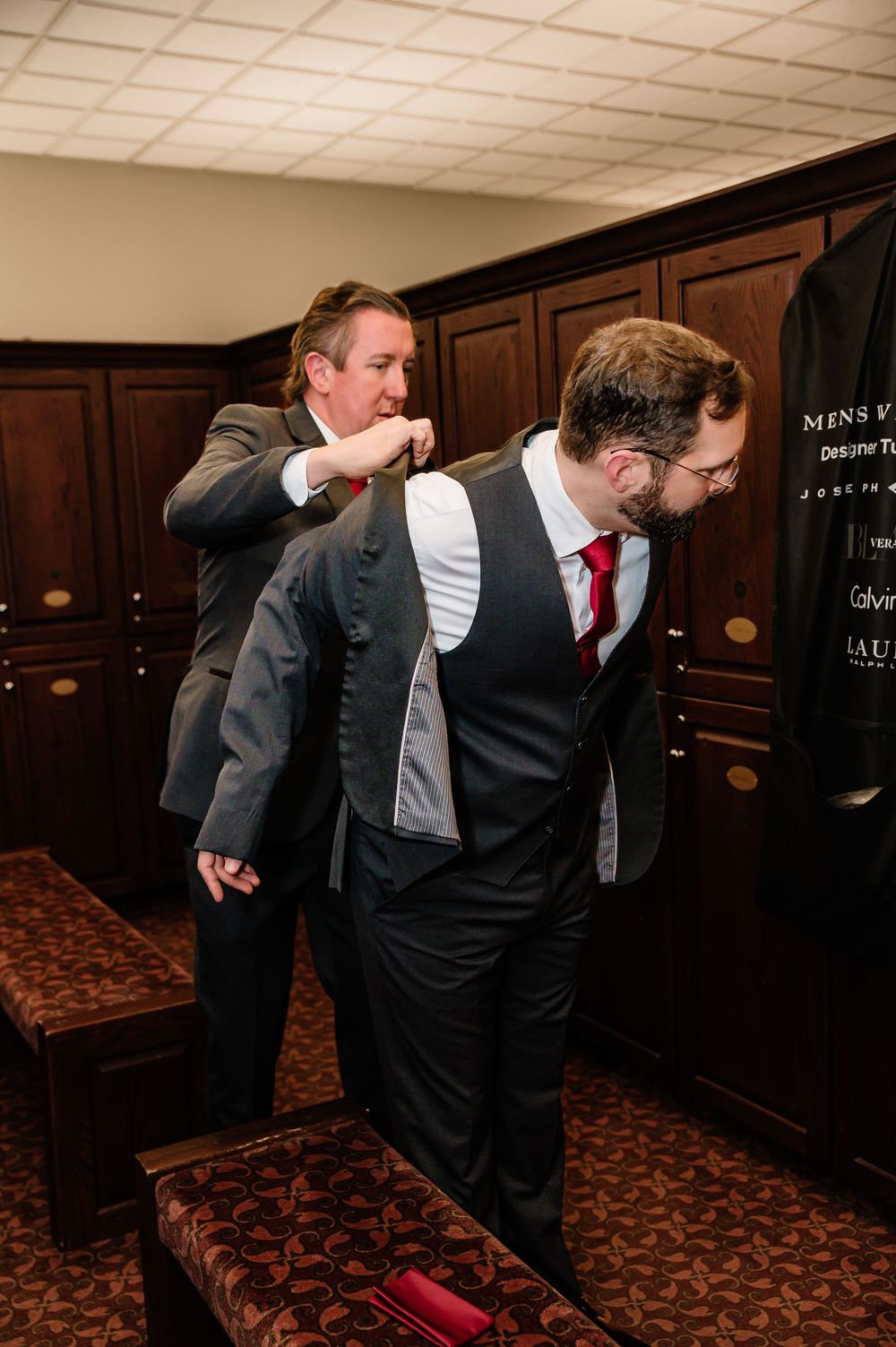 Two groomsmen in dark suits and red ties help each other dress in a wood-paneled fitting room.