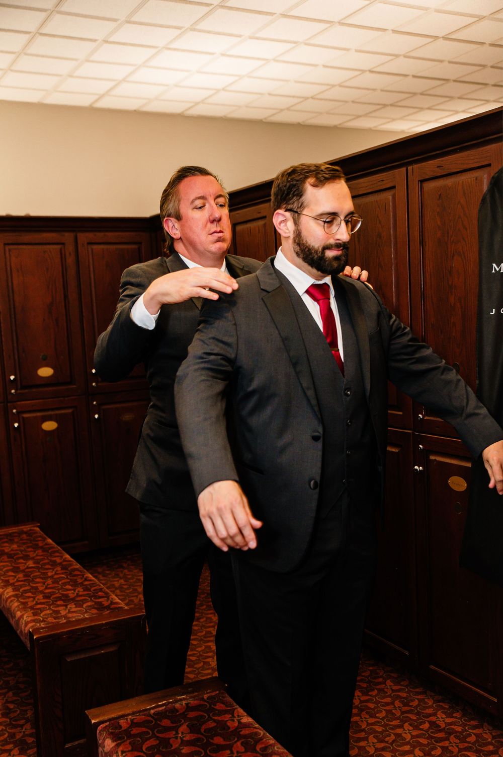 Two men in dark suits prepare in a wood-paneled room, one helping adjust the other's jacket before a formal event.