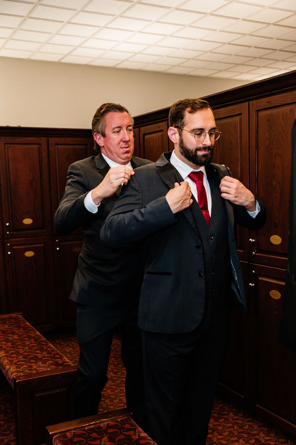 Two men in dark suits with red ties prepare in a wood-paneled room, one adjusting the other's jacket collar.