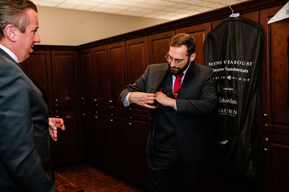 Two men in suits inside a formal menswear store, one adjusting his jacket near a garment bag.