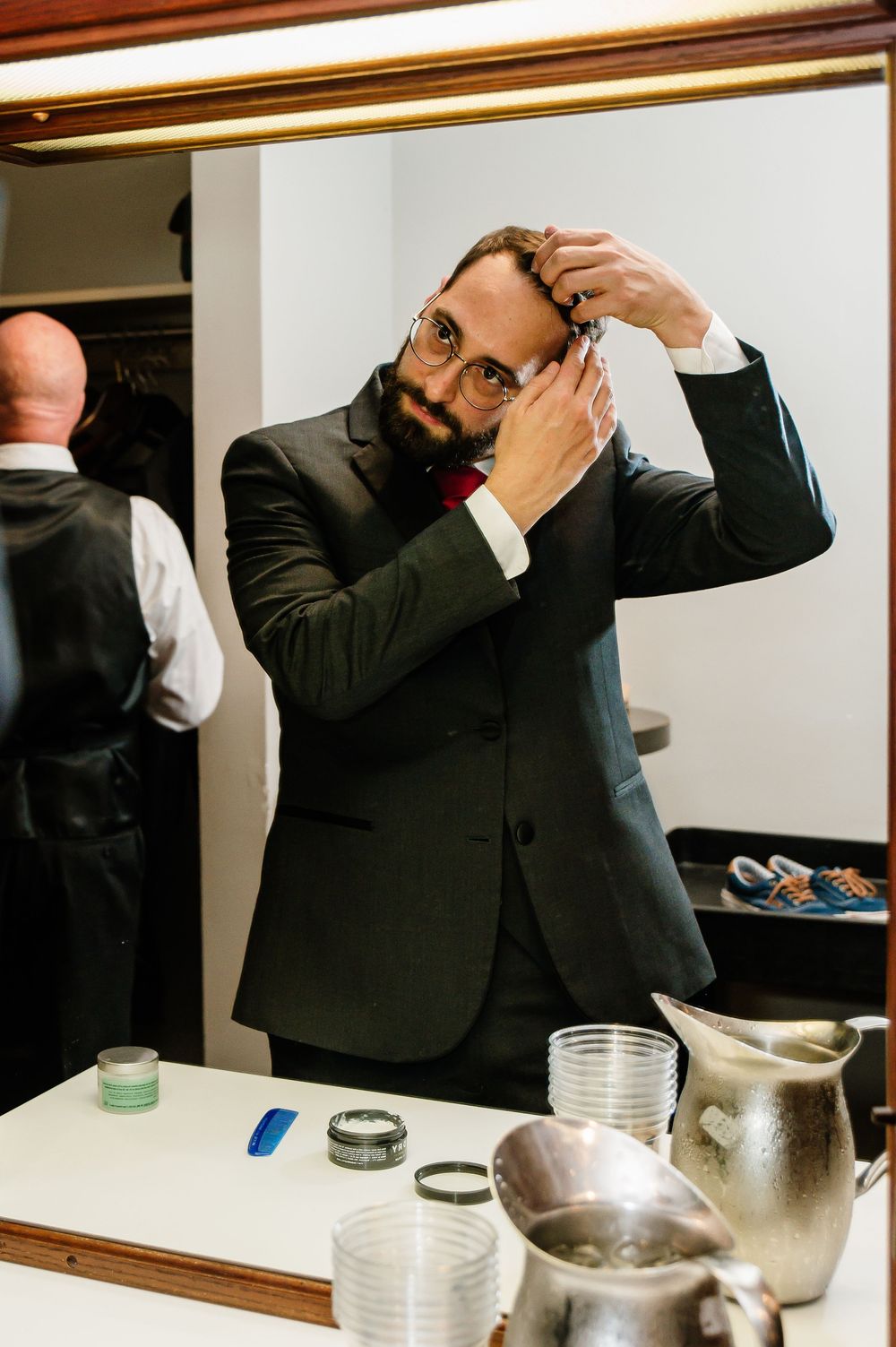 Bearded man in dark suit adjusting hair while looking in mirror, with glasses and red bow tie.
