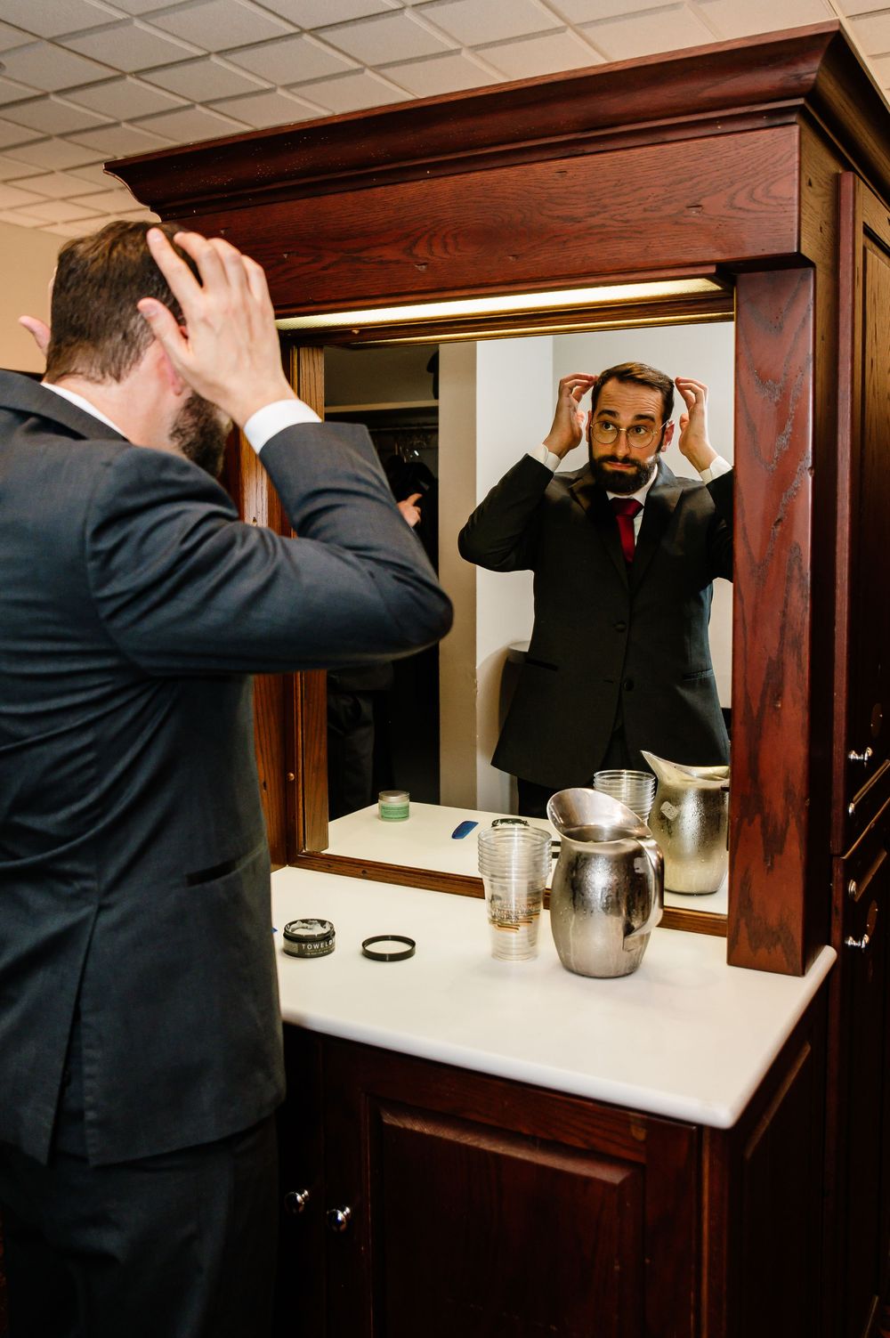 A well-dressed man in a dark suit adjusts his hair in a wooden framed bathroom mirror before a formal event.