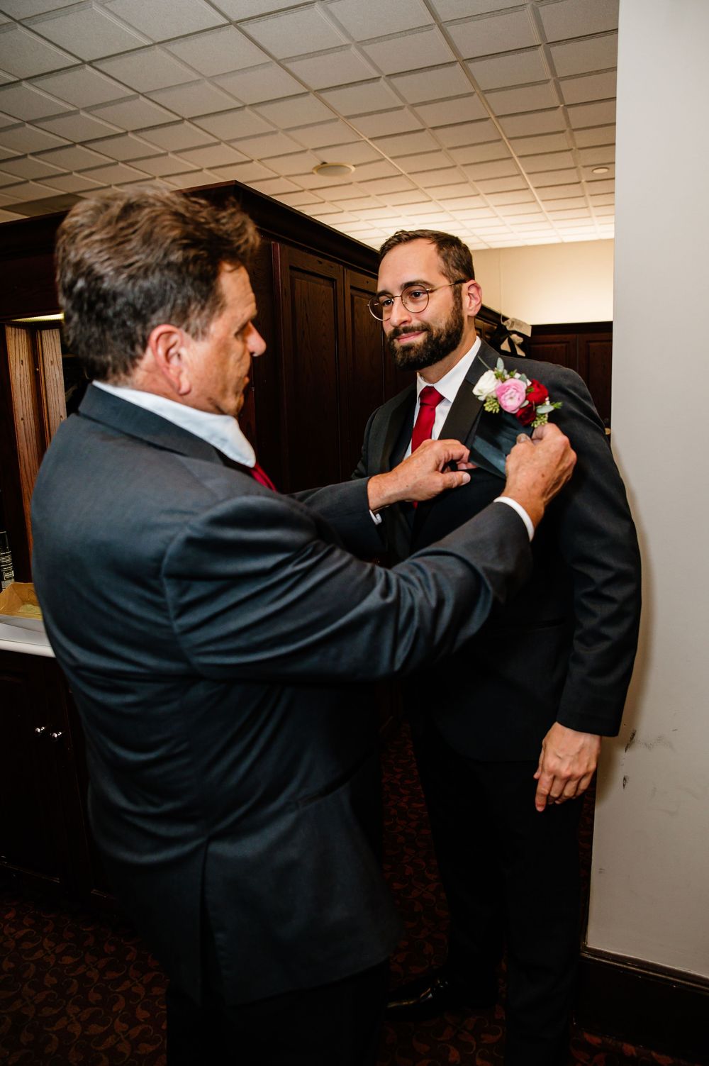 A man pins a boutonniere on a groom in a dark suit before a wedding ceremony.
