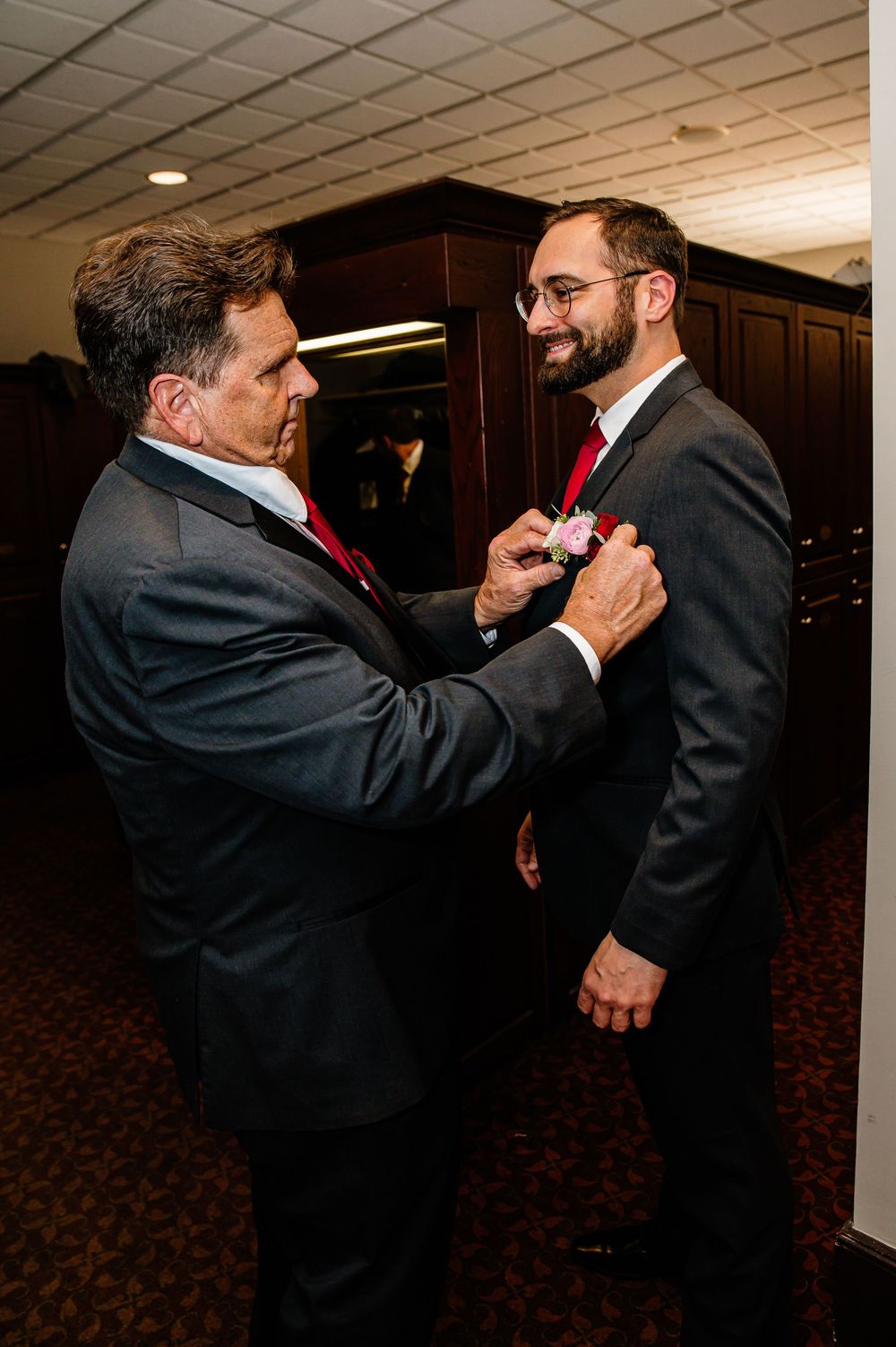 A man pins a boutonniere on a groom in a dark suit with red tie before a wedding ceremony.