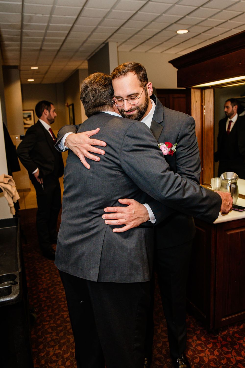 Two men in gray suits share a warm hug at a wedding reception, one wearing glasses and a boutonniere.