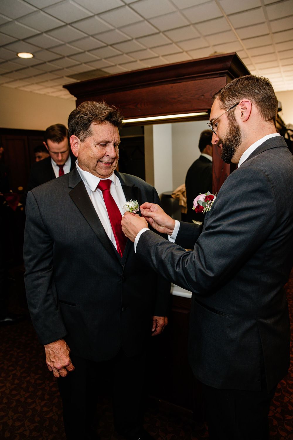 A bearded groom pins a boutonniere on an older man in a black tuxedo with red tie at a wedding.