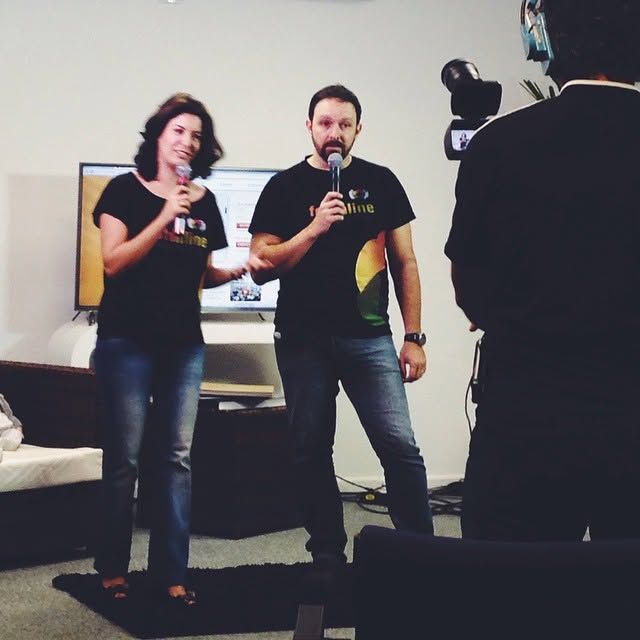 A man and woman in black shirts hold microphones while presenting indoors, filmed by a camera operator.