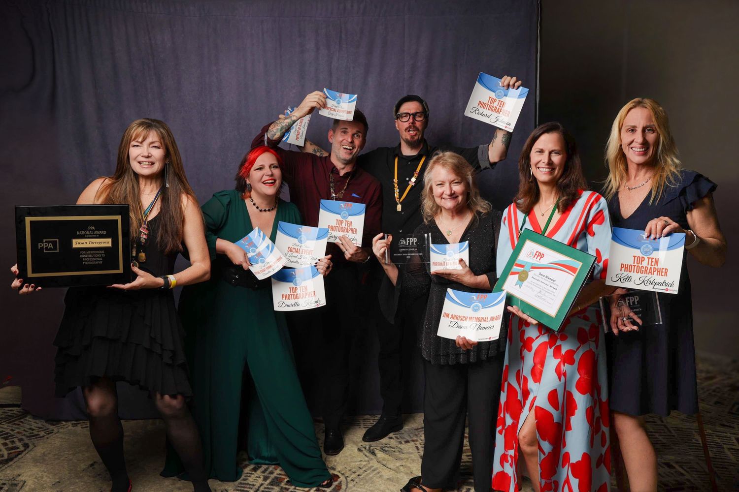 Group of seven people smiling and holding awards and certificates at an indoor recognition ceremony.
