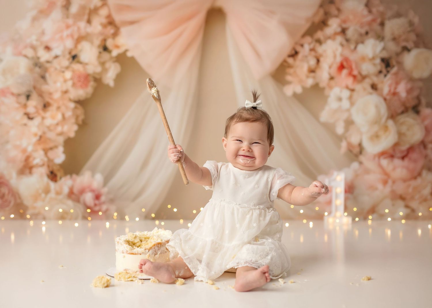 Baby girl in white dress smiles at first birthday cake smash with pink floral backdrop and fairy lights.