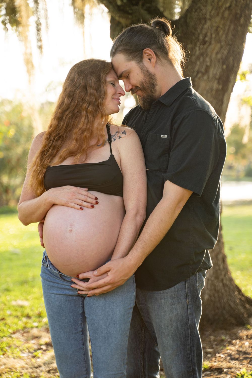 Couple sharing a tender moment outdoors, man embracing pregnant woman's baby bump near a sunlit tree.