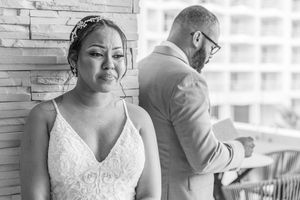 Black and white photo of a bride in a lace gown standing back-to-back with a groom in a suit during a first look.