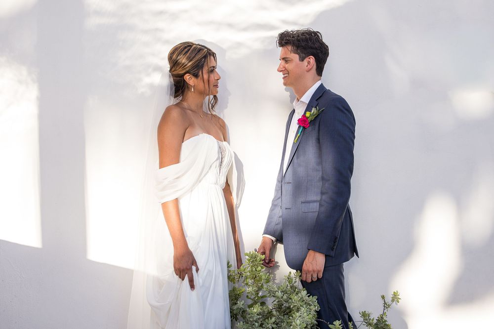 Bride in white off-shoulder gown and groom in navy suit share a romantic moment against a white wall.