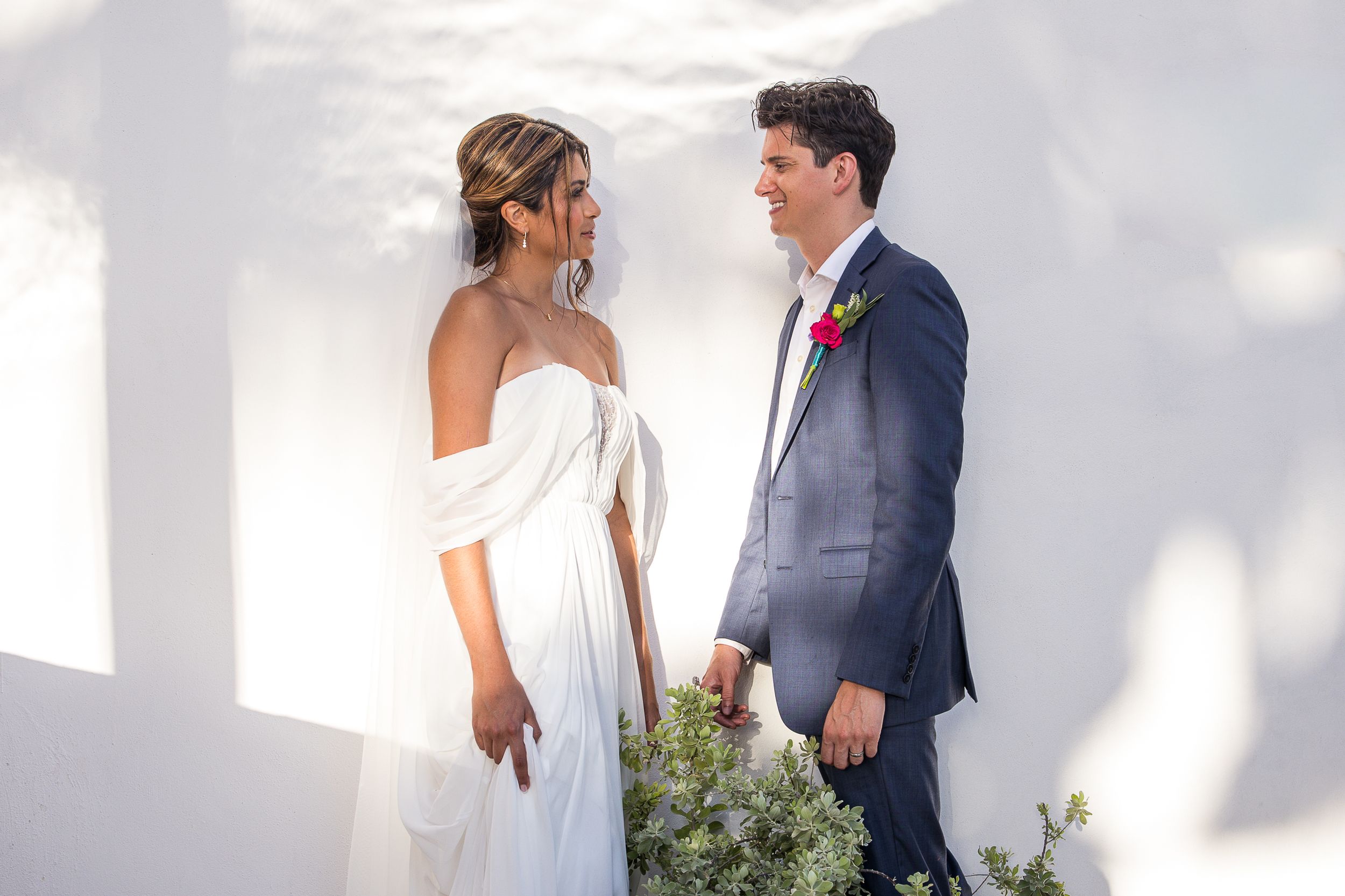 Bride in white off-shoulder gown and groom in navy suit share a romantic moment against a white wall.