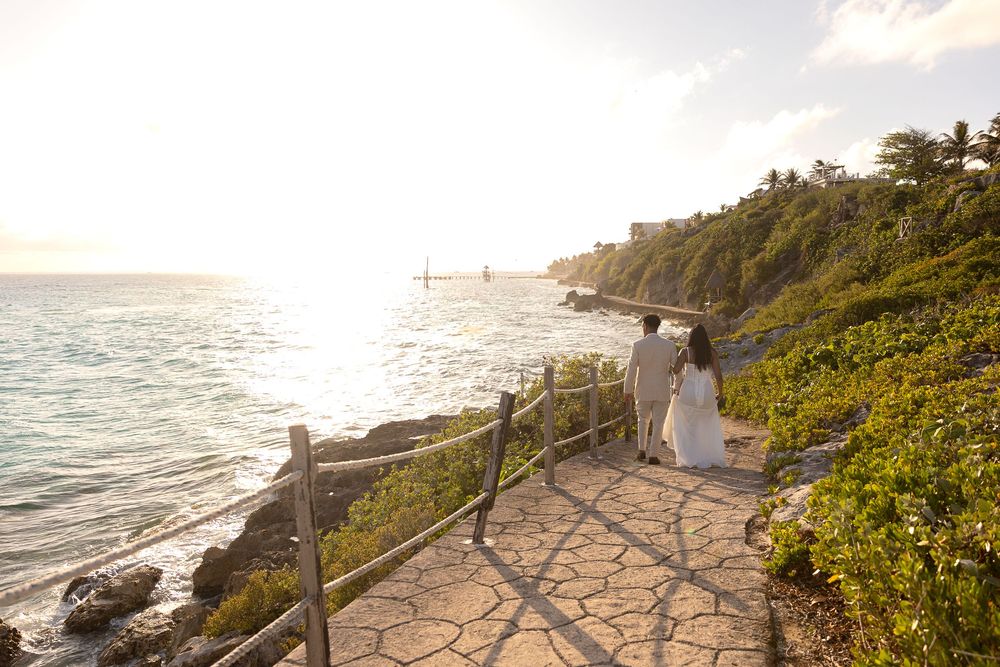 Couple in wedding attire walking along a scenic coastal path at sunset with ocean views and lush greenery.