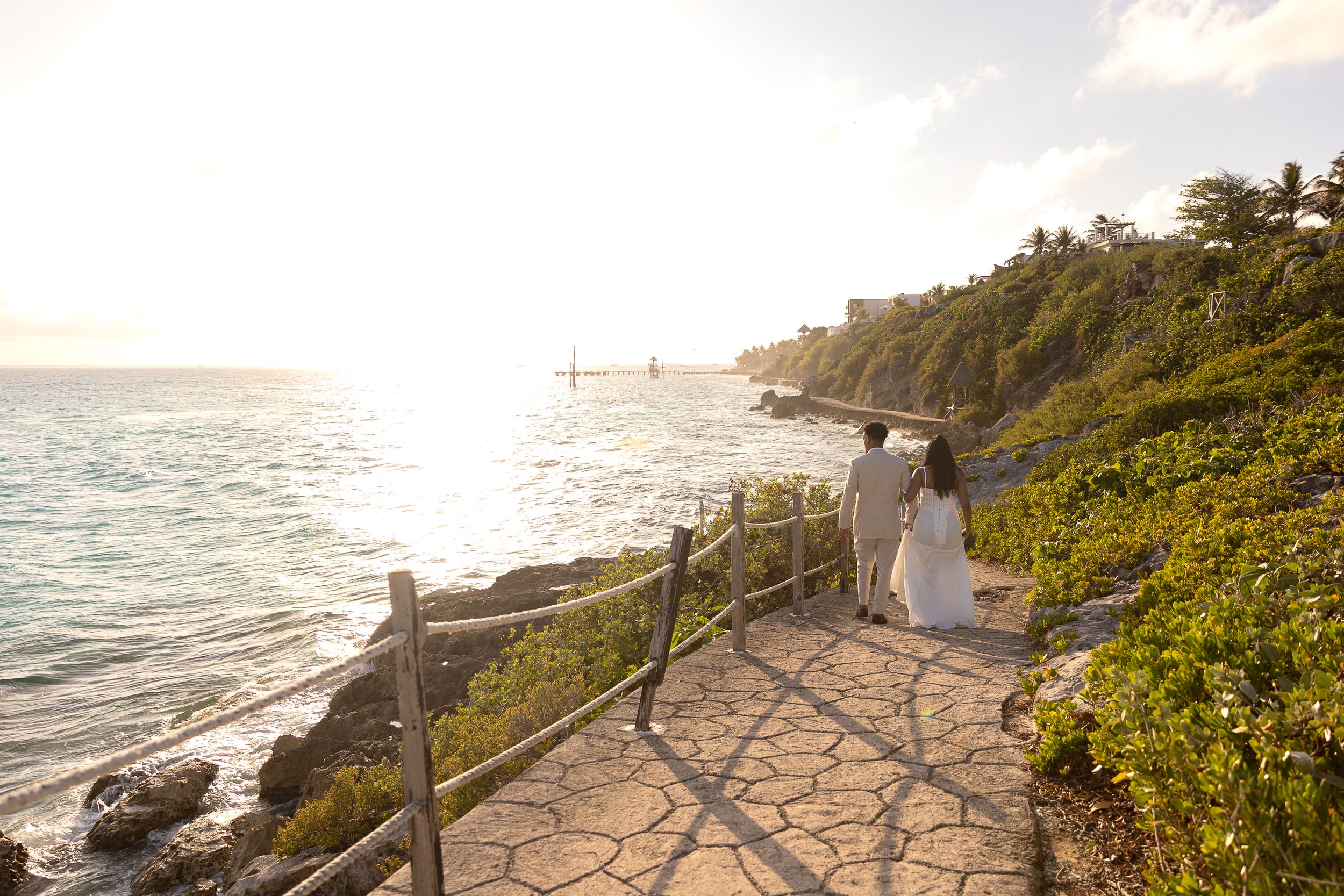 Couple in wedding attire walking along a scenic coastal path at sunset with ocean views and lush greenery.