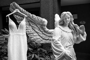 Angel statue holding a pearl-beaded wedding dress on hanger in black and white garden photo.