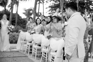 Groom in white suit watches bride walk down aisle at outdoor wedding ceremony with guests seated in white chairs.