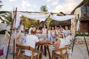 Outdoor beach wedding ceremony under a white draped chuppah with guests in white attire, palm trees and ocean in background.