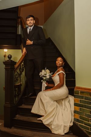 Interracial couple in wedding attire posing on ornate wooden staircase, bride in white gown holding bouquet.