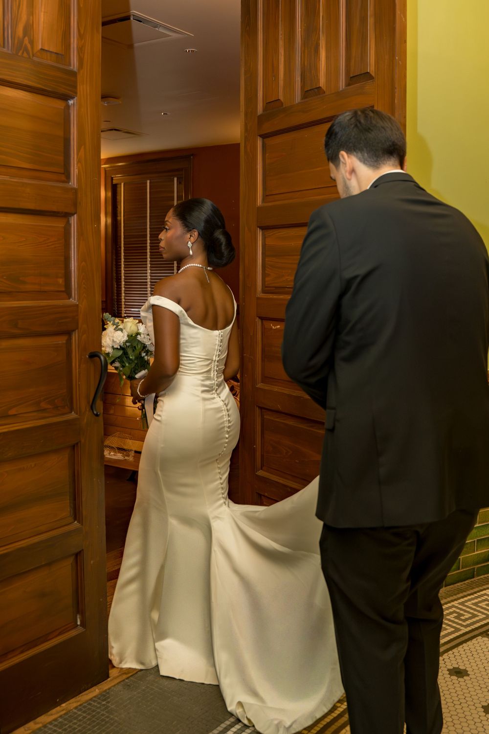 Bride in white gown holding bouquet with groom in black suit entering wooden doors at wedding venue.