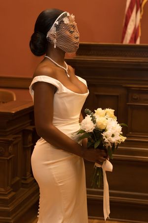 Elegant bride in white off-shoulder gown with birdcage veil holds yellow and white bouquet in ornate venue.