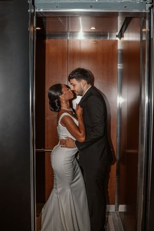 A bride and groom share a romantic moment in a wood-paneled elevator, dressed in elegant wedding attire.