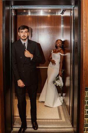 Interracial couple in wedding attire posing in elevator, groom in black suit, bride in white mermaid gown holding bouquet.