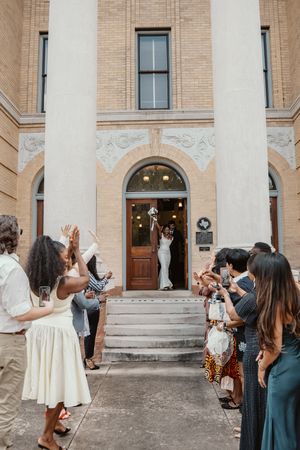 Newlyweds exit a historic brick church as guests cheer and clap along a pathway in a joyful wedding send-off.