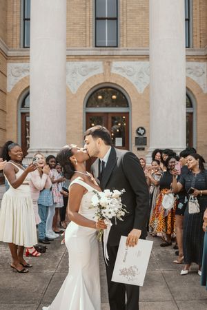 Newlywed couple sharing a kiss outside a historic brick church while guests celebrate their wedding ceremony.