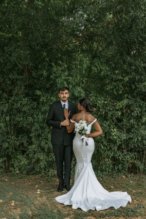 Interracial couple posing in wedding attire before lush green trees, bride in white mermaid gown holding bouquet.