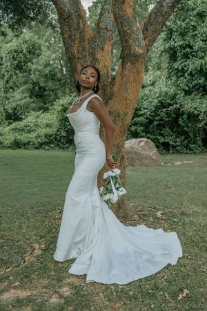 Elegant bride in white mermaid gown with train poses by a tree in lush green garden, holding a bouquet.