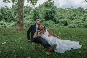 Bride and groom sitting on lush green grass in a park, with the bride leaning against her groom in wedding attire.