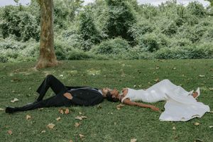 Bride and groom lying on green grass in a park, wearing wedding attire, surrounded by lush trees and autumn leaves.
