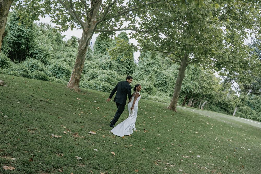 A bride and groom walk hand in hand on a lush green hillside surrounded by tall trees on their wedding day.