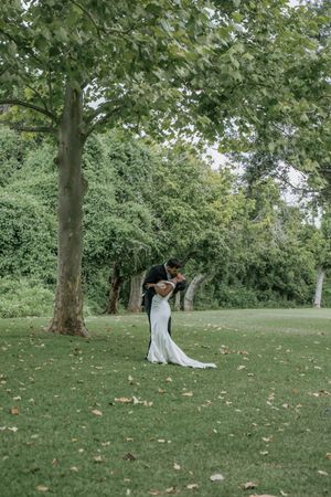 A bride and groom share a romantic embrace on a lush green lawn surrounded by tall trees.