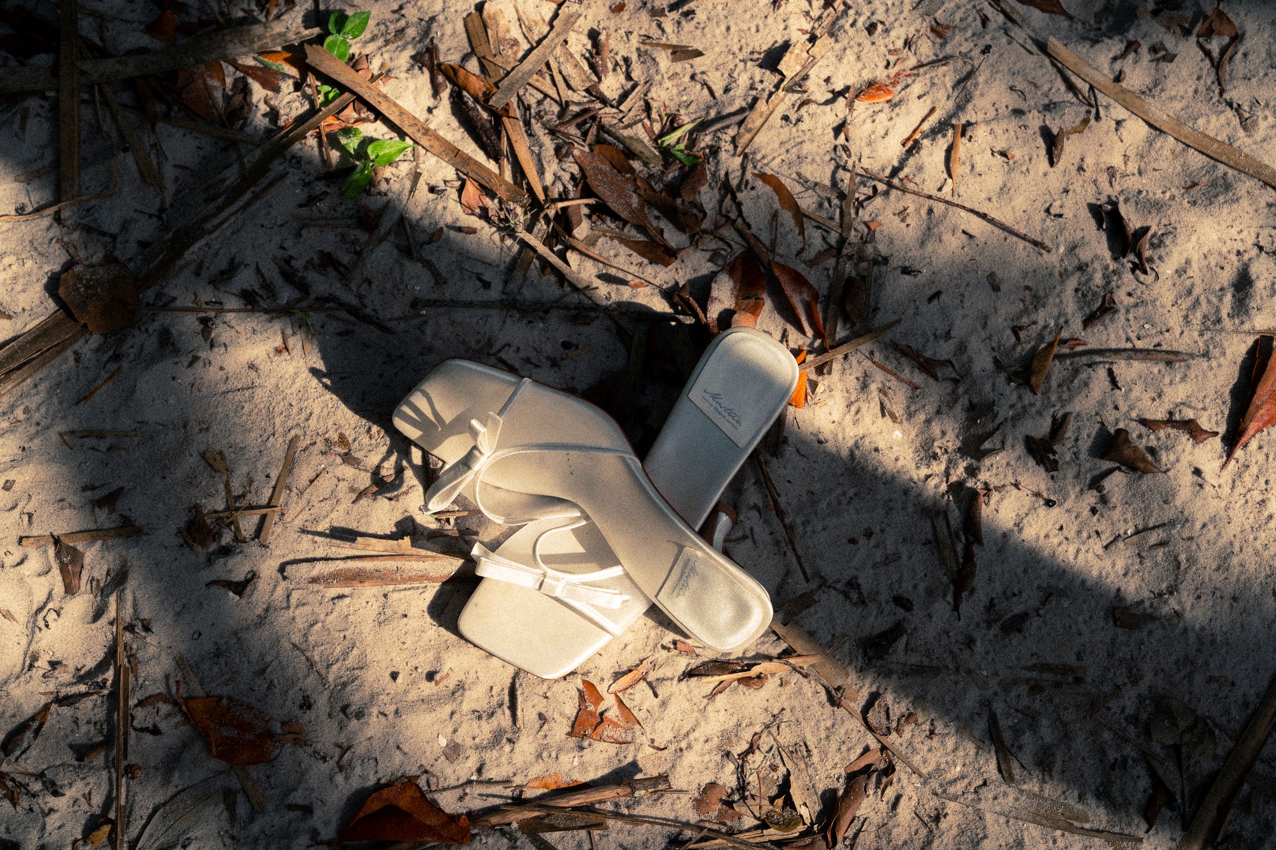 A discarded white flip-flop sandal lies abandoned on sandy ground surrounded by dry leaves and debris.