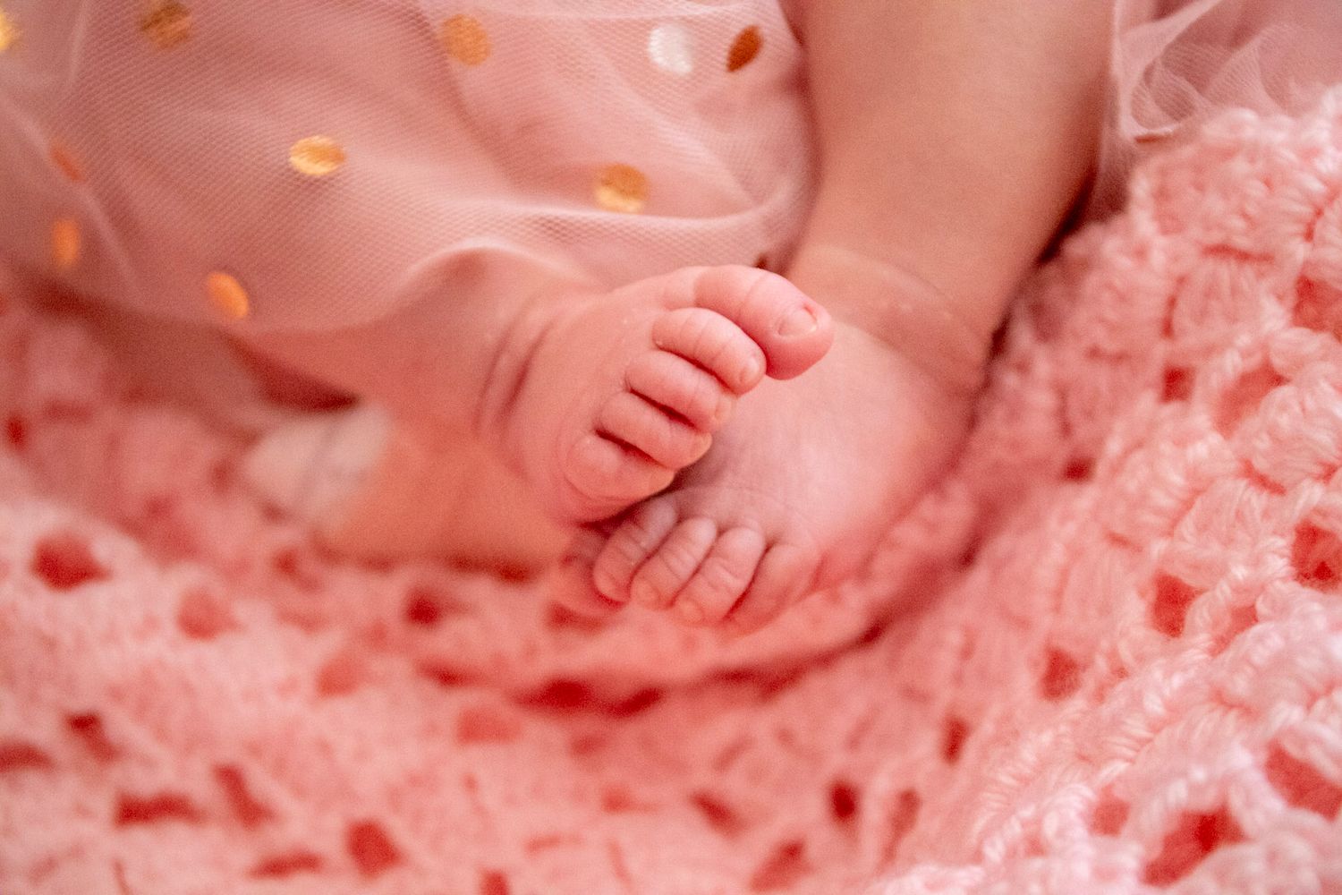 Close-up of tiny newborn baby feet resting on a soft pink knitted blanket with a polka dot dress.
