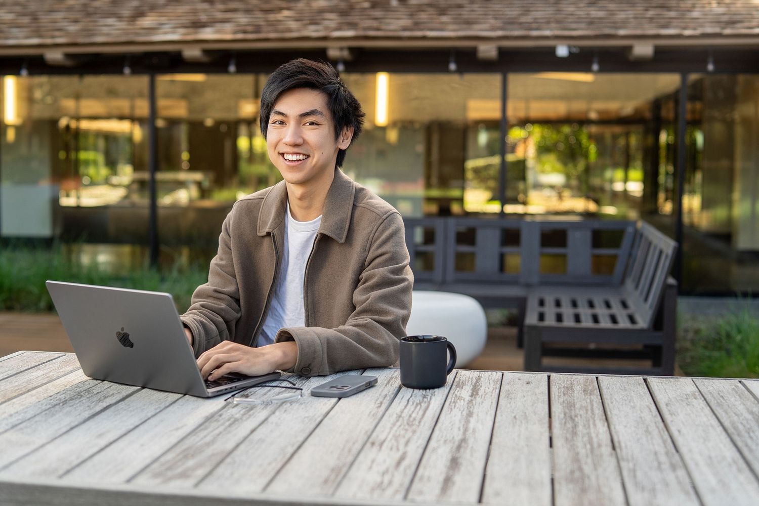 Young Asian man smiling while working on MacBook laptop at outdoor wooden table near modern building.