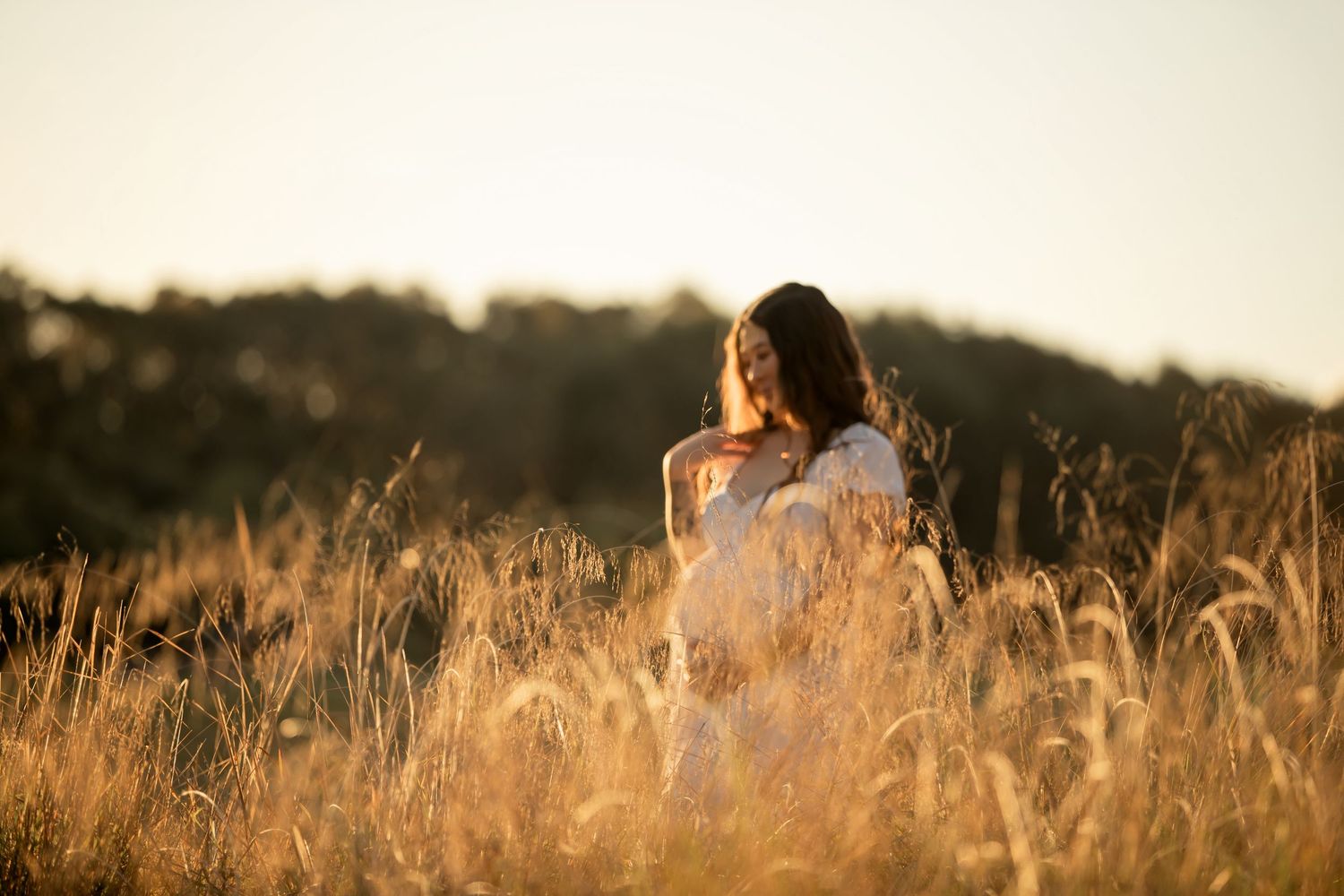 A young woman in a white dress stands in golden wheat fields during sunset, bathed in warm glowing light.