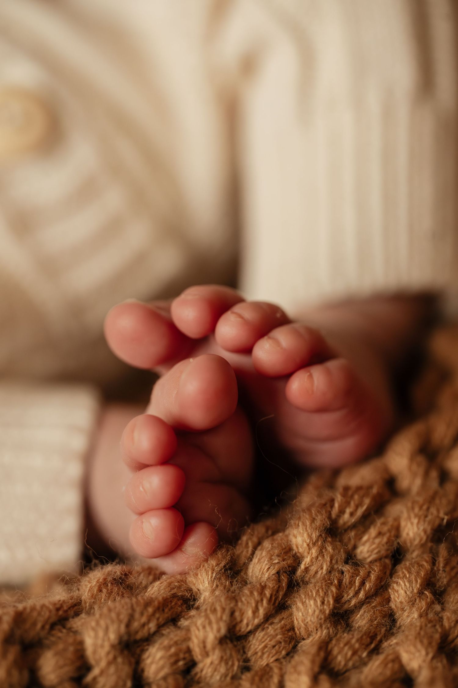 Close-up of tiny newborn baby feet resting on a soft brown knit blanket, wearing a cream knit outfit.