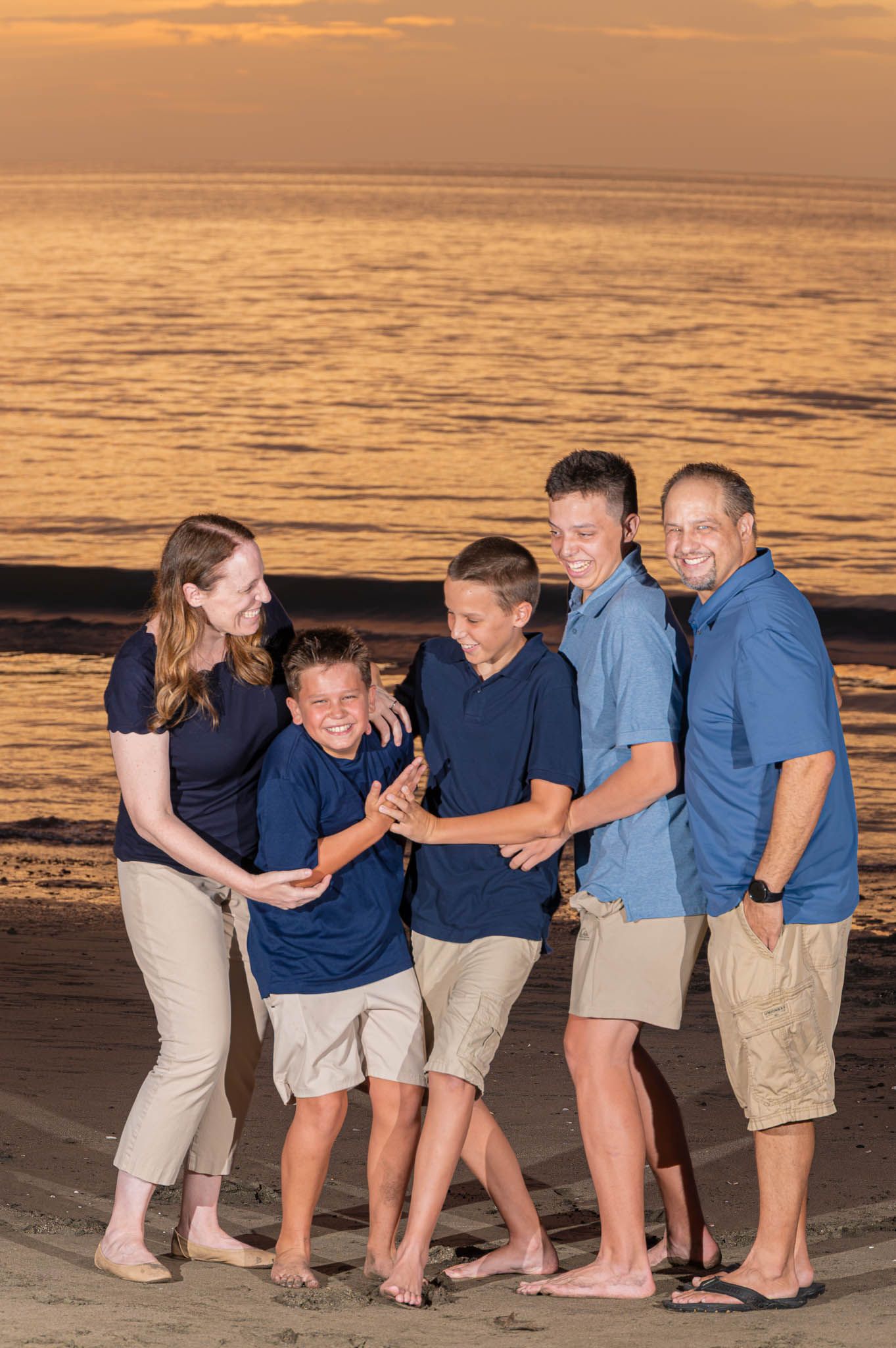 Family of five laughing together on a beach at golden sunset, wearing coordinating navy and khaki outfits.