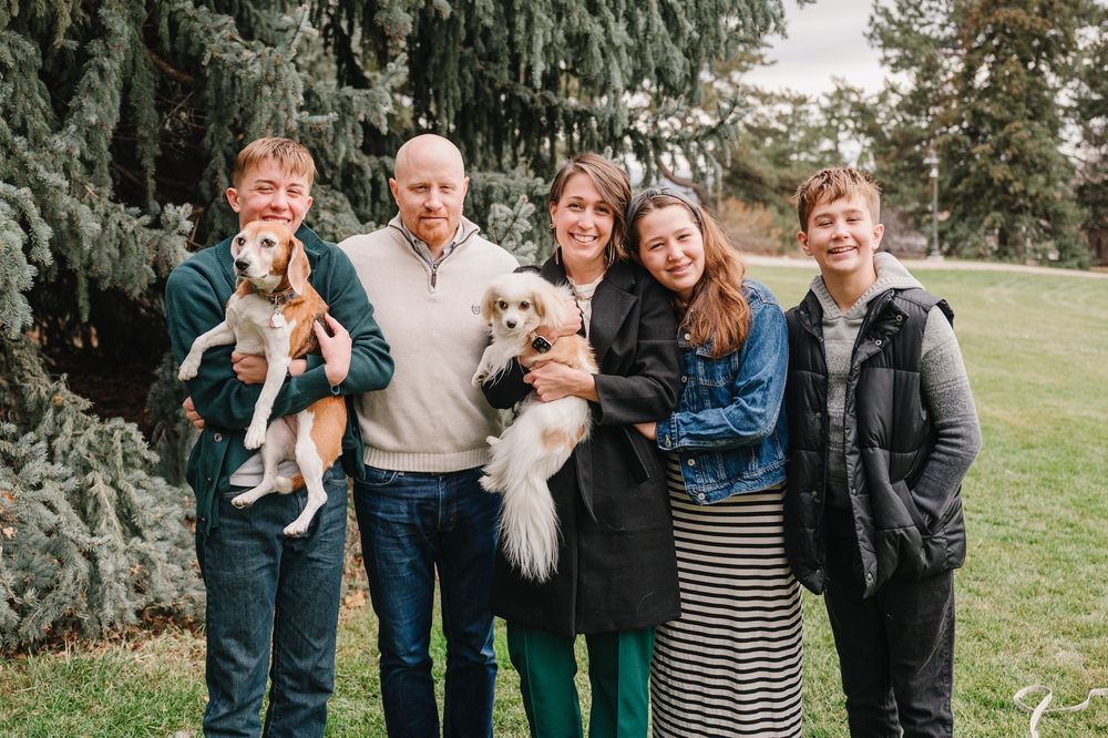 Family of five smiling outdoors with two dogs, posing near evergreen trees on a cloudy day.