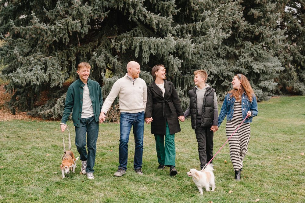 Family of five walking dogs in a park with evergreen trees, smiling and holding hands outdoors.