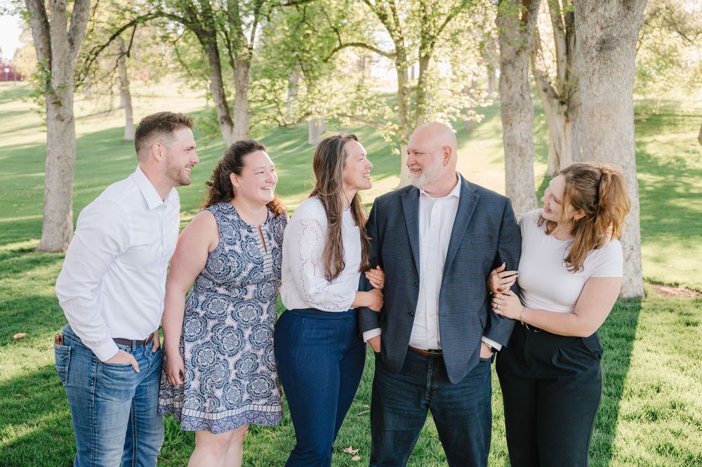 A group of five people laughing together while standing outdoors in a sunlit park with trees in the background.