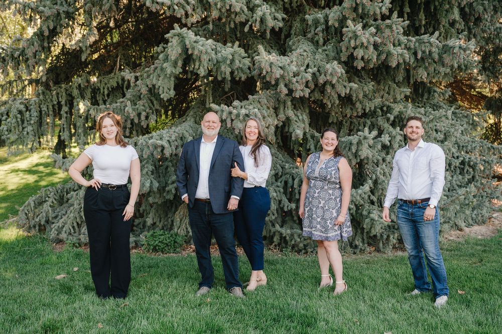 A group of five people pose together on a sunny day in front of a large evergreen tree on a grassy lawn.