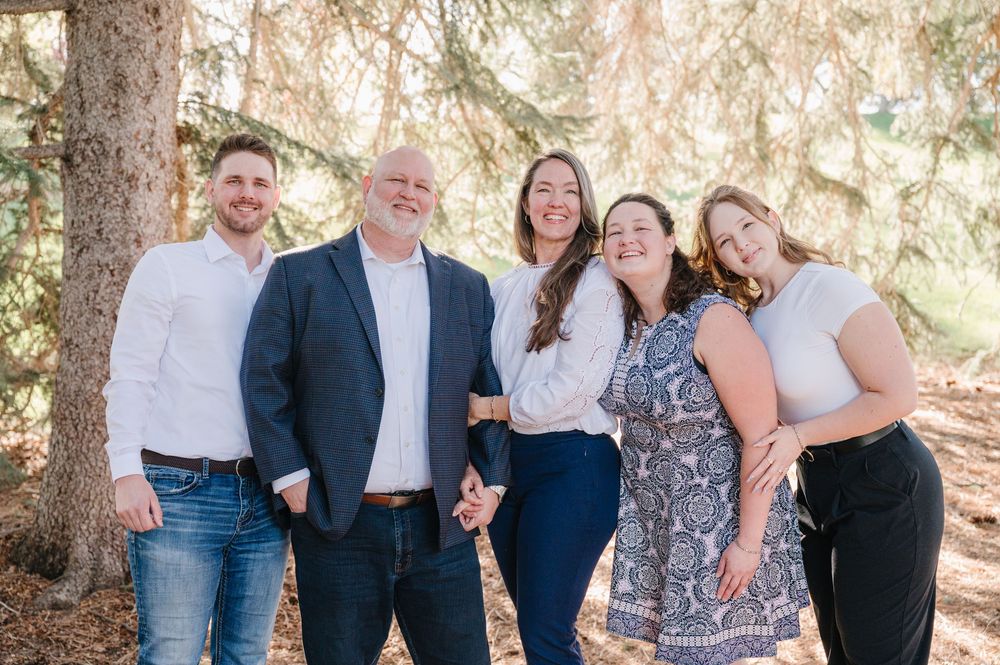 Five people stand together smiling outdoors under evergreen trees in a casual family portrait setting.
