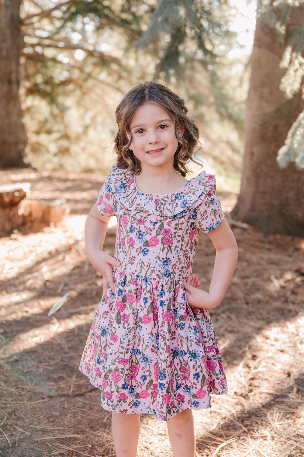 Young girl in pink floral dress posing outdoors among trees in warm sunlight.
